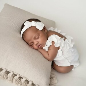 Sleeping newborn baby resting on a beige tassel pillow, dressed in a timeless ivory newborn girl outfit. The organic cotton fabric drapes softly, creating a peaceful, luminous baby announcement lifestyle portrait.