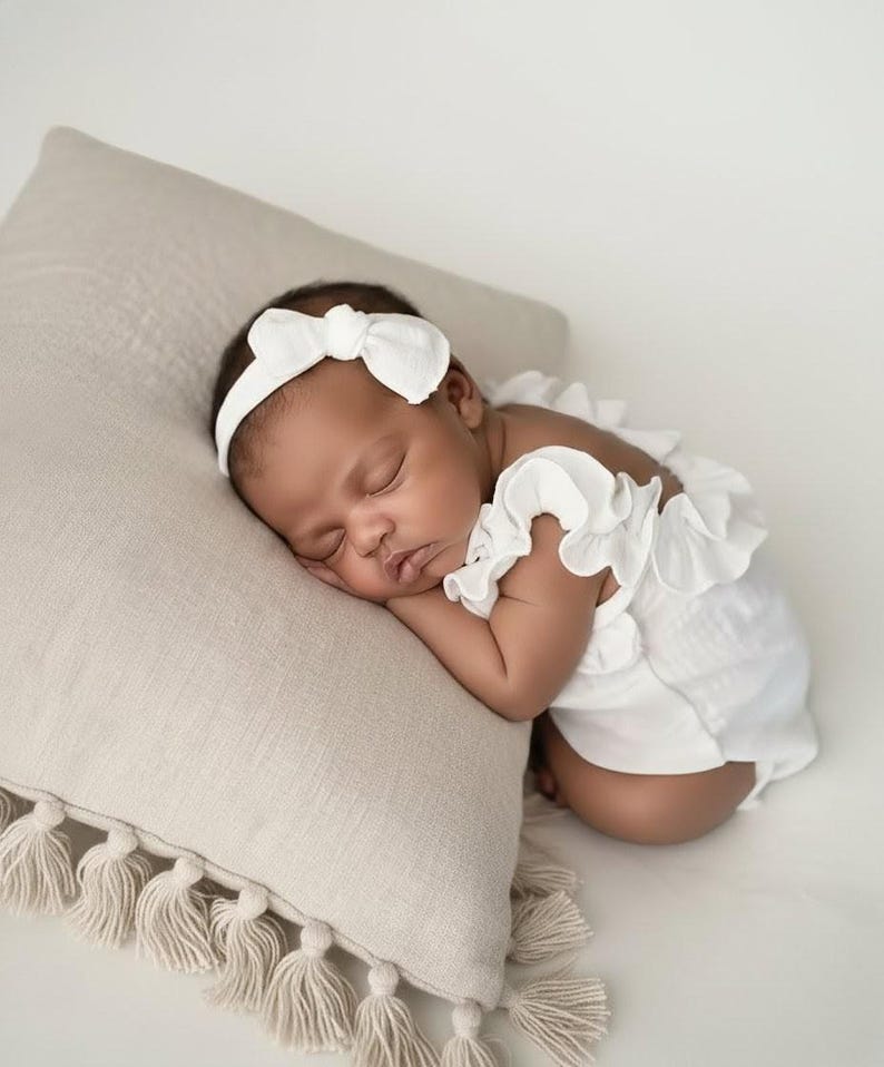 Sleeping newborn baby resting on a beige tassel pillow, dressed in a timeless ivory newborn girl outfit. The organic cotton fabric drapes softly, creating a peaceful, luminous baby announcement lifestyle portrait.