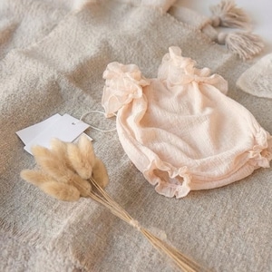 Close-up macro shot of a delicate newborn romper revealing the crinkle cotton fabric and high-stretch leg cuffs. The romantic oversized frill collar makes this a timeless baby shower gift, styled minimally with dried bunny tail grass.