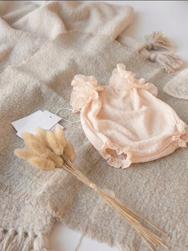Close-up macro shot of a delicate newborn romper revealing the crinkle cotton fabric and high-stretch leg cuffs. The romantic oversized frill collar makes this a timeless baby shower gift, styled minimally with dried bunny tail grass.