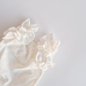 Detailed crop of organic cotton baby romper sleeve ruffles against a stark white background. The intricate hand-pleated ivory fabric casts soft shadows, demonstrating the high-quality craftsmanship of this newborn gift.