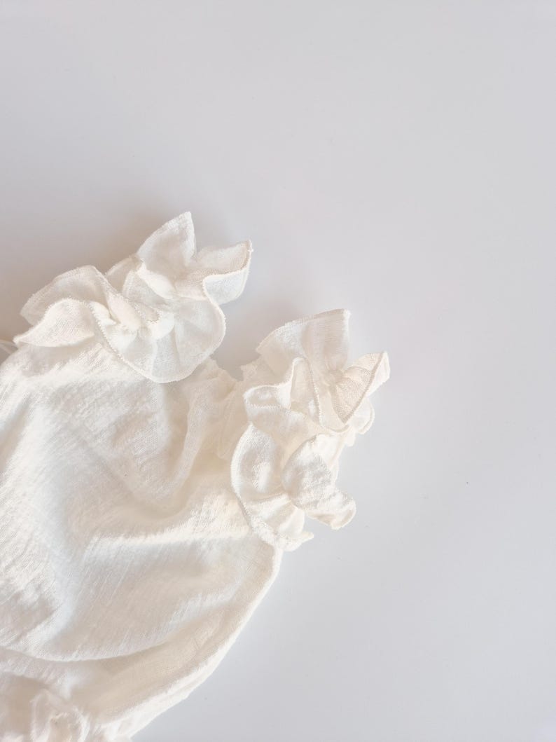 Detailed crop of organic cotton baby romper sleeve ruffles against a stark white background. The intricate hand-pleated ivory fabric casts soft shadows, demonstrating the high-quality craftsmanship of this newborn gift.