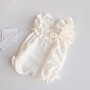 Ivory newborn outfit flat lay featuring a smocked bodice and voluminous ruffle neckline. Matte cotton texture visible against a white background with brand tags. Ideal minimalistic baby shower gift presentation.