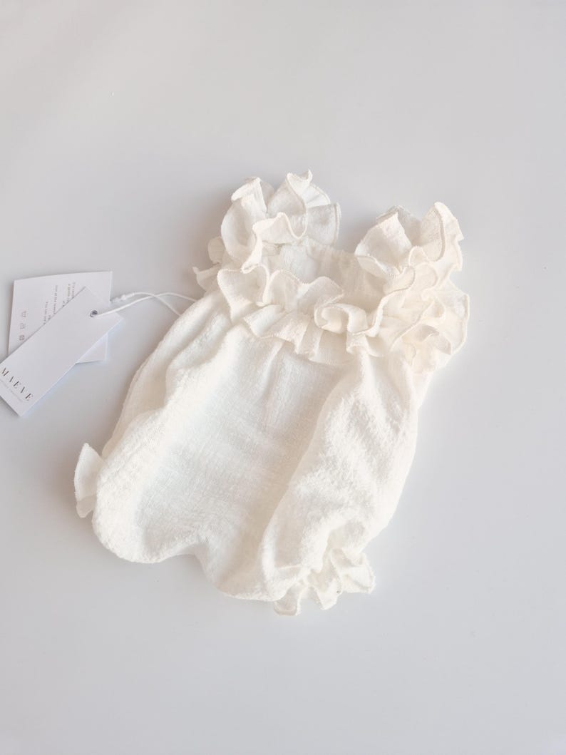 Ivory newborn outfit flat lay featuring a smocked bodice and voluminous ruffle neckline. Matte cotton texture visible against a white background with brand tags. Ideal minimalistic baby shower gift presentation.