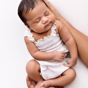 May include: A newborn infant with dark hair sleeping peacefully while wearing a white sleeveless romper. The garment features a smocked bodice with a textured fabric and delicate ruffled trim along the shoulder straps and neckline.