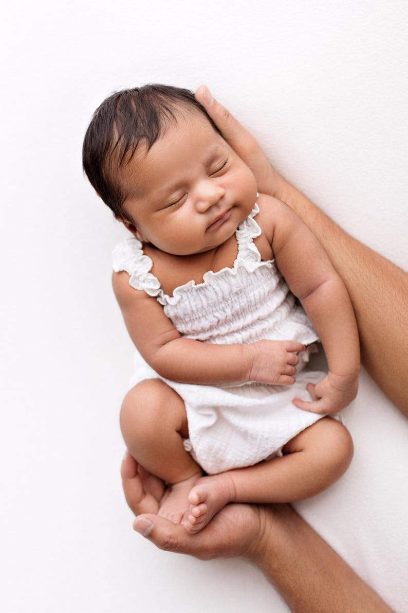 May include: A newborn infant with dark hair sleeping peacefully while wearing a white sleeveless romper. The garment features a smocked bodice with a textured fabric and delicate ruffled trim along the shoulder straps and neckline.