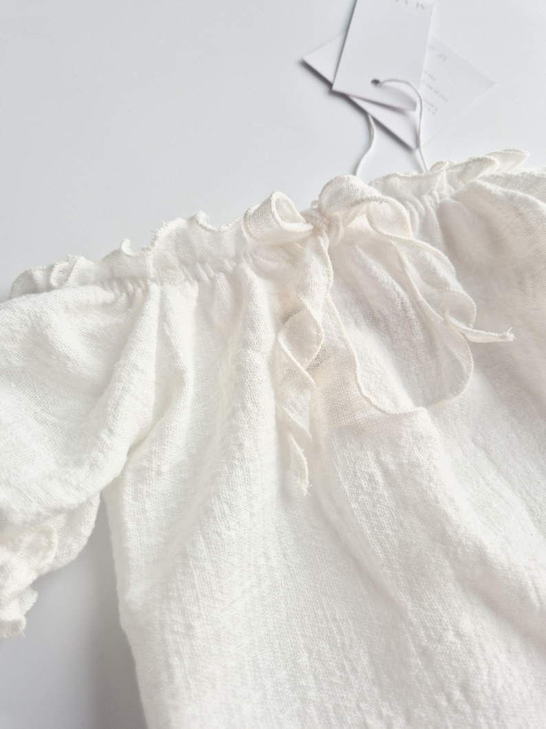Detailed view of an elegant tie-back closure on a pure white textured baby outfit. The soft crinkle cotton and delicate bow add a touch of vintage charm, creating a heartwarming coming home outfit and versatile newborn photo prop.