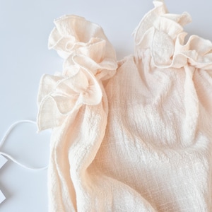 Soft, nostalgic glow of off-white cotton fabric. Close-up on the intricate, draped ruffle detailing of a creamy vanilla baby romper. A highly textured photography prop essential for fine art newborn studio aesthetics.