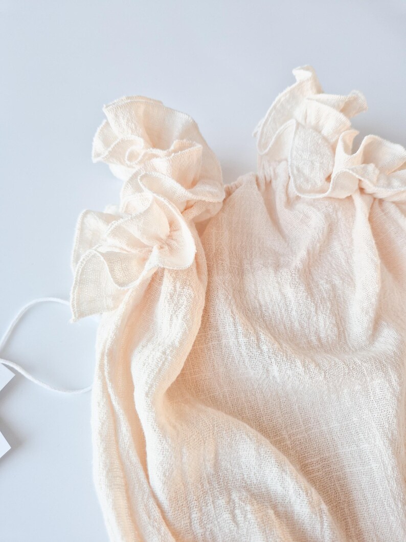 Soft, nostalgic glow of off-white cotton fabric. Close-up on the intricate, draped ruffle detailing of a creamy vanilla baby romper. A highly textured photography prop essential for fine art newborn studio aesthetics.