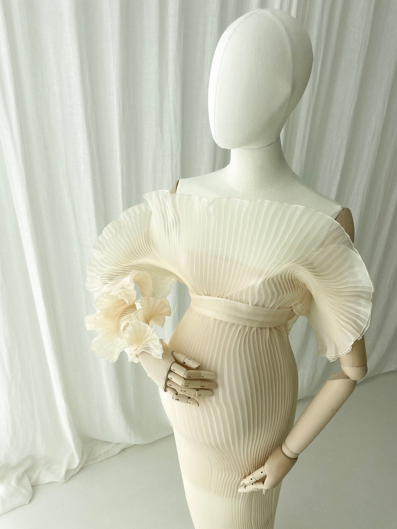 Mannequin detail shot showcasing the voluminous, sculptural ruffle sleeve of the Celessia gown. The warm peach color of the finely pleated fabric is ideal for complementary golden hour outdoor photography.