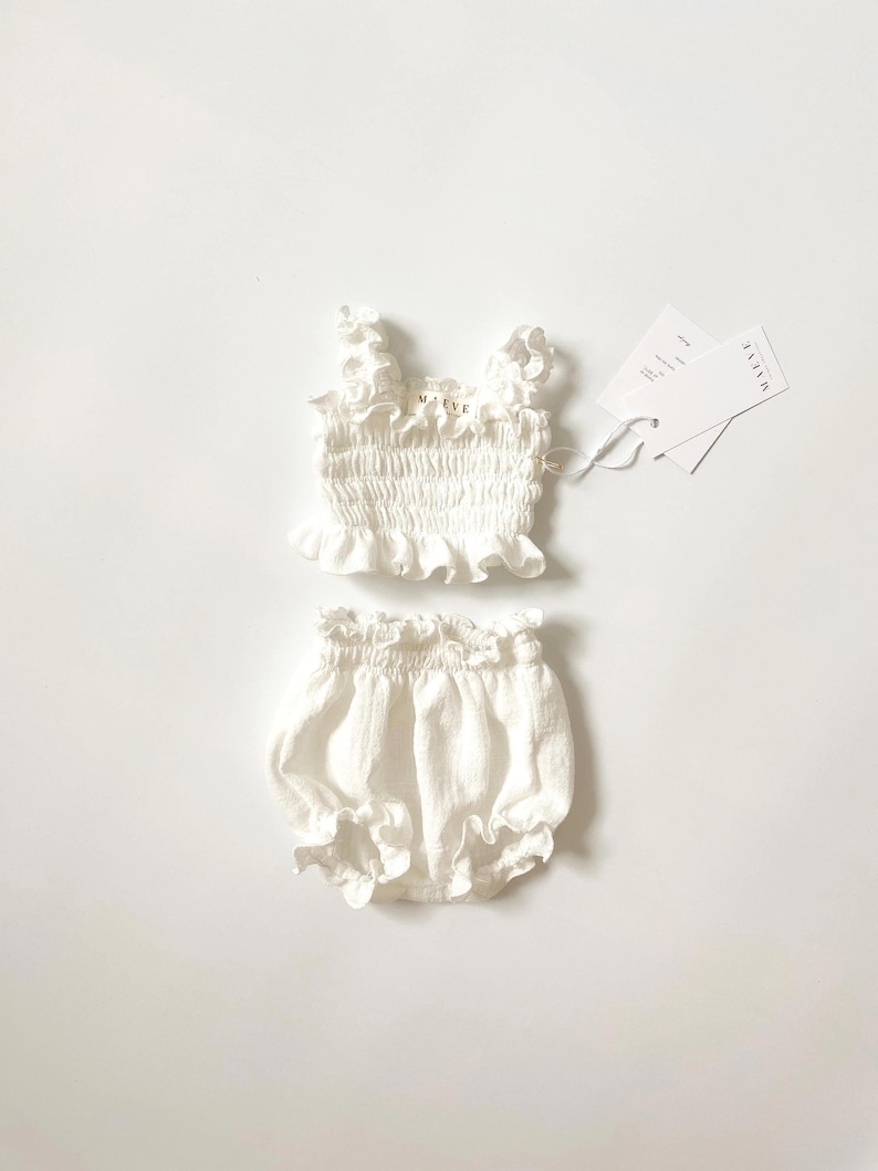 May include: A white two-piece set consisting of a sleeveless top and bloomers. The top features a smocked bodice, ruffled straps, and a ruffled hem. The bloomers have an elasticized waistband and leg openings with ruffles. Tags labeled MAEVE are attached.