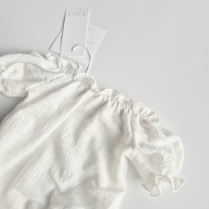 Flat lay close-up of a white baby romper showcasing breathable crinkle cotton and delicate puff sleeves. This highly textured photography prop offers a zero-prep, organic foundation for boho baby clothes styling in studio settings.
