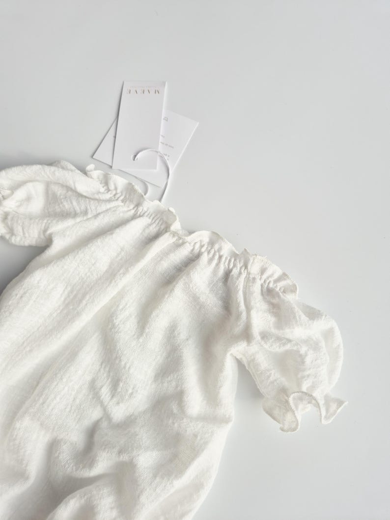 Flat lay close-up of a white baby romper showcasing breathable crinkle cotton and delicate puff sleeves. This highly textured photography prop offers a zero-prep, organic foundation for boho baby clothes styling in studio settings.