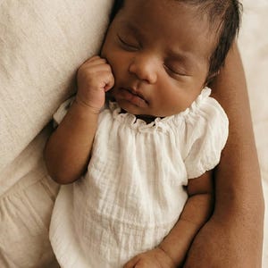 Close-up of a peaceful baby in adult arms wearing a white cotton romper styled with a classic boatneck silhouette. The gentle neckline and breathable texture make this practical piece a heartwarming coming home outfit.