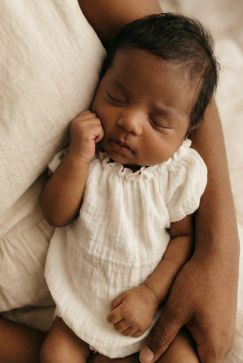 Close-up of a peaceful baby in adult arms wearing a white cotton romper styled with a classic boatneck silhouette. The gentle neckline and breathable texture make this practical piece a heartwarming coming home outfit.