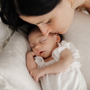 Mother lovingly kissing her sleeping baby who wears a comfortable, matte-finish ivory newborn romper. The non-restricting smocked design flatters sensitive skin, creating a flawless maternal-bond aesthetic for lifestyle sessions.