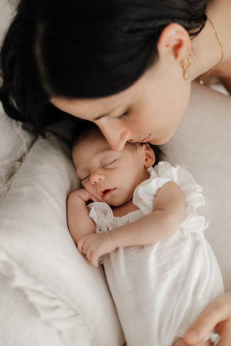 Mother lovingly kissing her sleeping baby who wears a comfortable, matte-finish ivory newborn romper. The non-restricting smocked design flatters sensitive skin, creating a flawless maternal-bond aesthetic for lifestyle sessions.