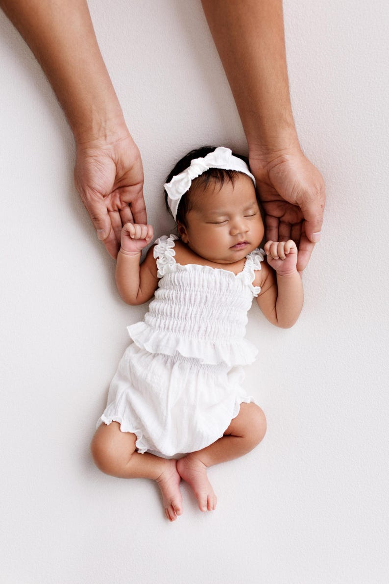 Sleeping infant in a white crinkled cotton two-piece newborn girl outfit. Adult hands gently frame the baby from above against a seamless studio backdrop. High-key lighting highlights the smocked texture of these ethereal newborn photo props.