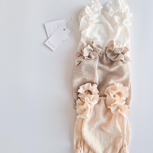 Flat lay of three textured cotton baby rompers in ivory, beige, and peach. Features high-volume pleated ruffles. Ideal neutral newborn props highlighting breathable organic fabric for fine art studio photography setups.