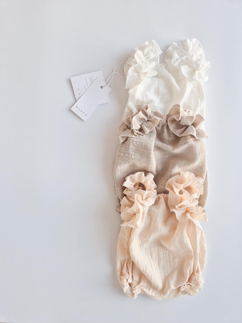 Flat lay of three textured cotton baby rompers in ivory, beige, and peach. Features high-volume pleated ruffles. Ideal neutral newborn props highlighting breathable organic fabric for fine art studio photography setups.