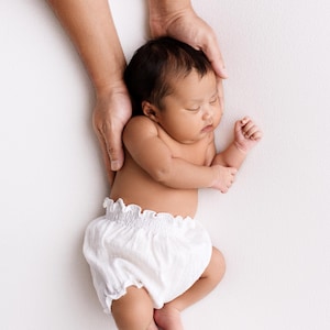 Top-down view of a sleeping newborn resting on a textured white backdrop, wearing only white light cotton puffy bloomers. Adult hands gently support the baby's back. Low-contrast lighting creates a soulful, minimalist newborn outfit portrait.