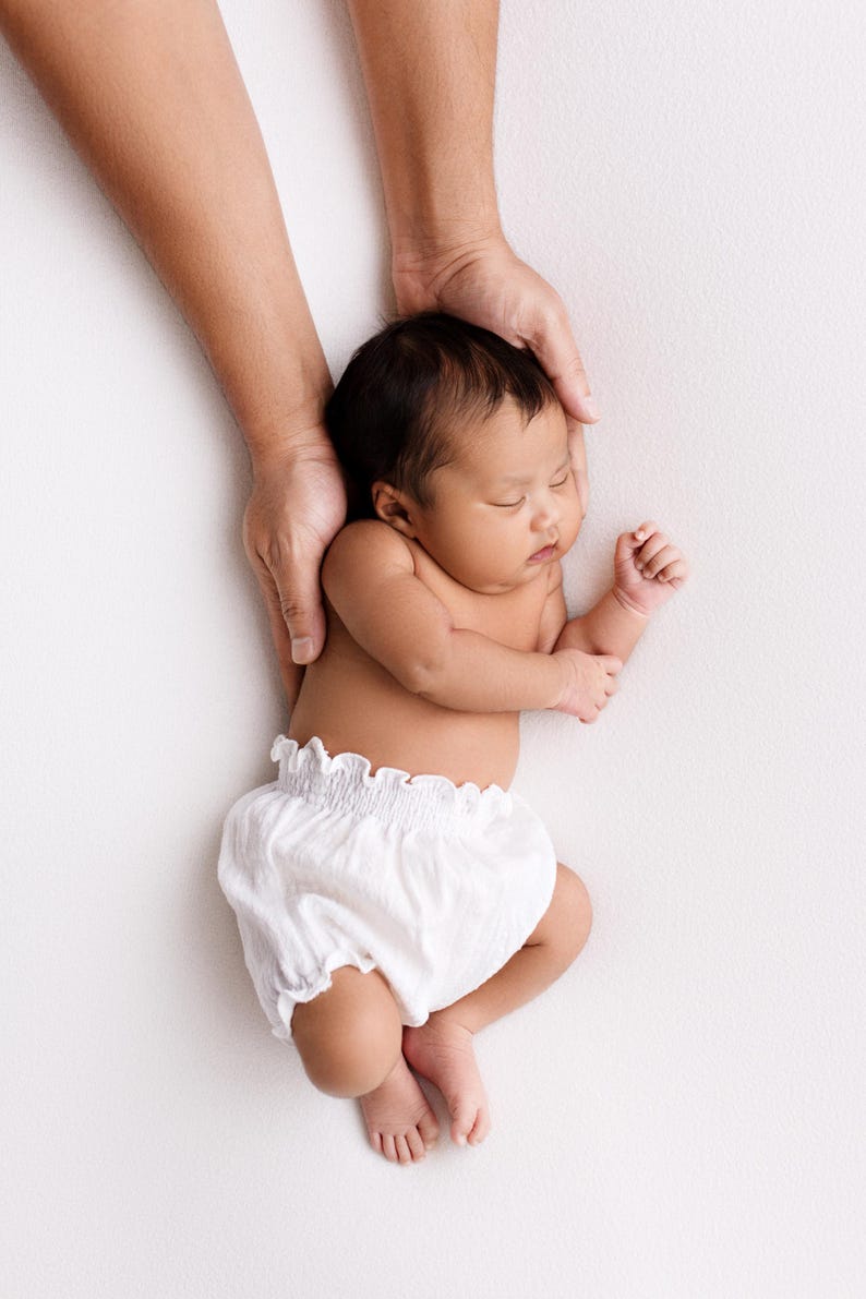 Top-down view of a sleeping newborn resting on a textured white backdrop, wearing only white light cotton puffy bloomers. Adult hands gently support the baby's back. Low-contrast lighting creates a soulful, minimalist newborn outfit portrait.