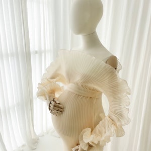 Profile view of the Celessia gown in Creamy Peach on a mannequin. The finely pleated fabric is fitted, highlighting the sculptural ruffle sleeve and the elegant mermaid flare at the hem.