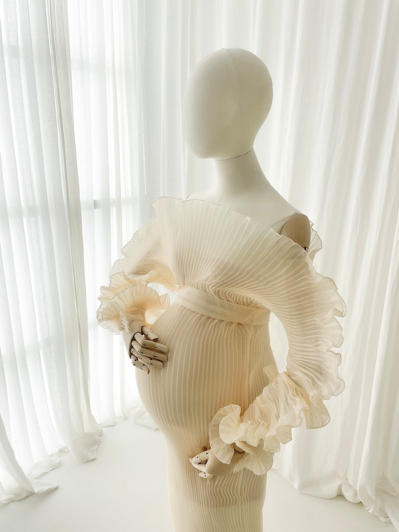 Profile view of the Celessia gown in Creamy Peach on a mannequin. The finely pleated fabric is fitted, highlighting the sculptural ruffle sleeve and the elegant mermaid flare at the hem.