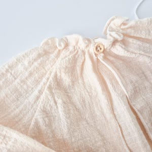 Detail shot of Lylou newborn girl outfit featuring a stretchy smocked elastic neckline for friction-free dressing. Buttery soft textured fabric in warm vanilla tone, ideal for cozy coming home apparel.