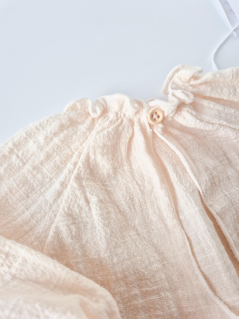 Detail shot of Lylou newborn girl outfit featuring a stretchy smocked elastic neckline for friction-free dressing. Buttery soft textured fabric in warm vanilla tone, ideal for cozy coming home apparel.