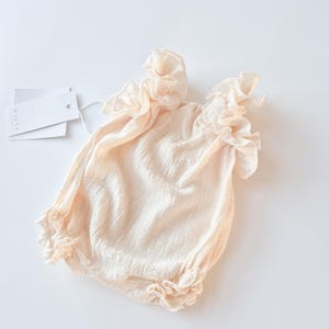 Full body flat lay of a newborn girl outfit in warm vanilla. Tactile, breathable textured cotton fabric with gentle elastic gathers at the leg. A delicate, zero-friction newborn photo prop for romantic baby styling.