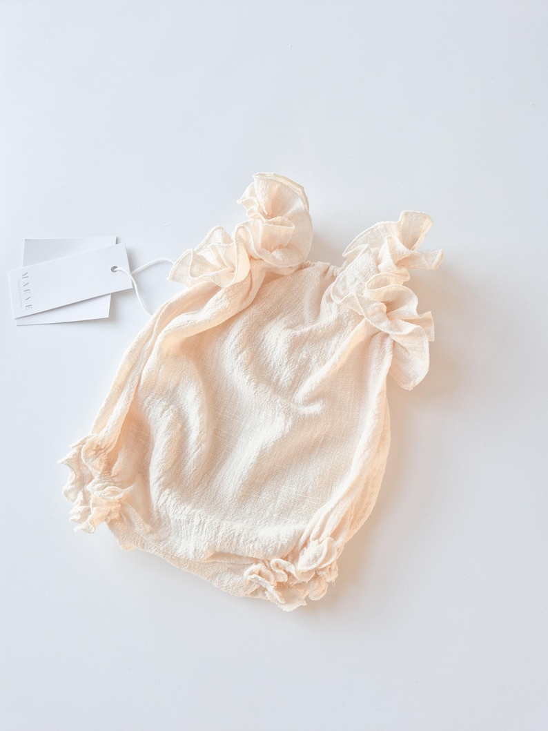 Full body flat lay of a newborn girl outfit in warm vanilla. Tactile, breathable textured cotton fabric with gentle elastic gathers at the leg. A delicate, zero-friction newborn photo prop for romantic baby styling.