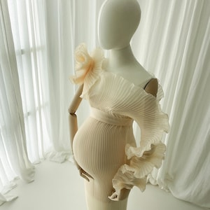 Side profile of the Celessia gown in soft apricot, styled on a mannequin. The sheer, light-catching pleated fabric sculpts the bump and emphasizes the dramatic off-the-shoulder ruffle accessory.