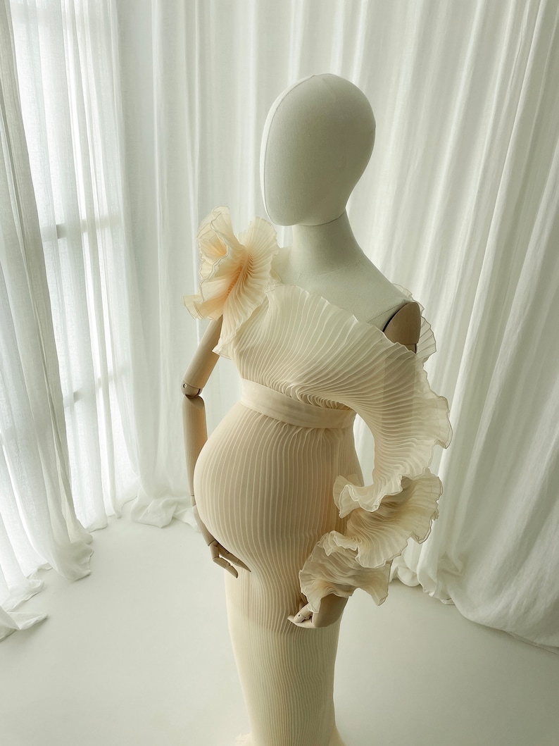 Side profile of the Celessia gown in soft apricot, styled on a mannequin. The sheer, light-catching pleated fabric sculpts the bump and emphasizes the dramatic off-the-shoulder ruffle accessory.