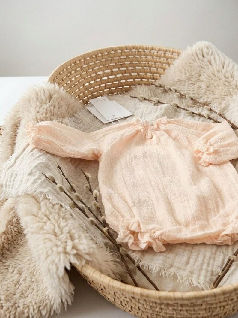 Close-up of the Lylou vanilla newborn romper resting in a basket. The premium 100% breathable crinkled cotton reveals 3D depth and buttery softness. Detailed shot of the smocked baby clothes texture.
