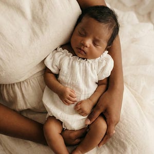 A tranquil newborn rests in an adult's arms, wearing a white crinkle cotton romper with puff sleeves. Designed to pair with maternity gowns, this cohesive mommy and me outfit creates a flawless, high-end aesthetic for lifestyle portraits.