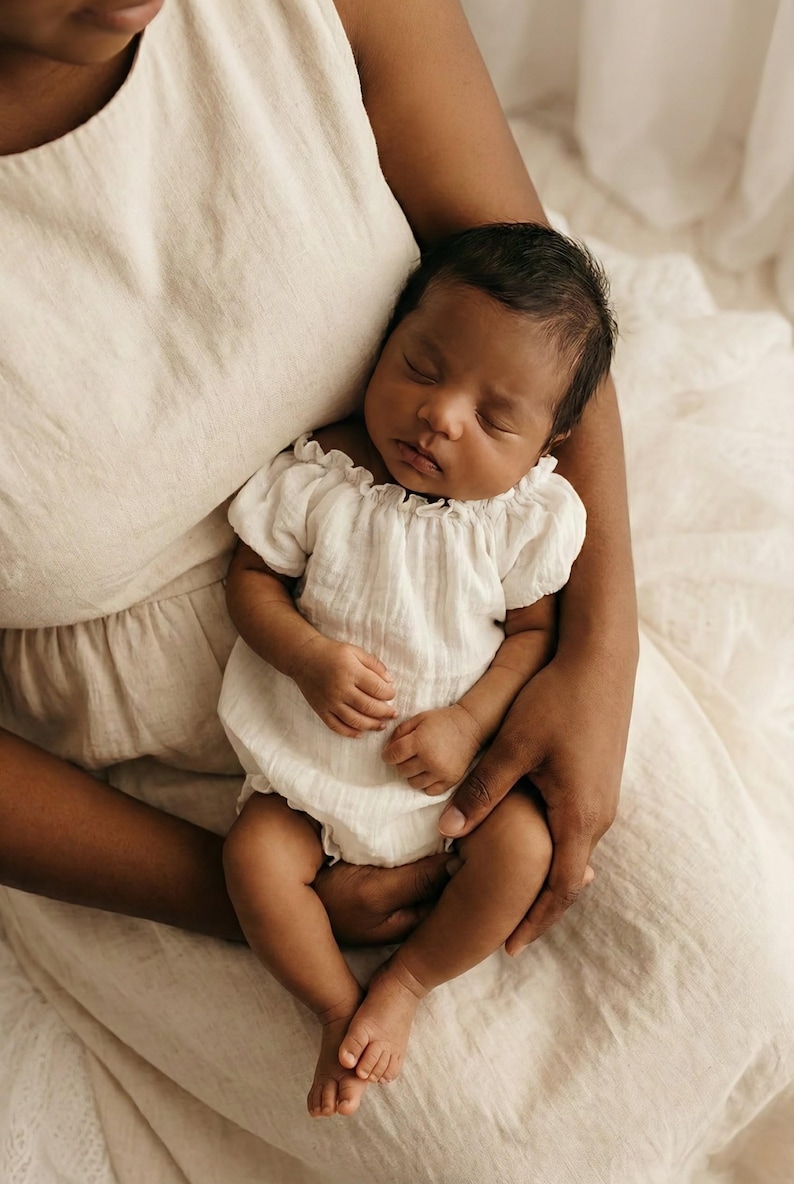 A tranquil newborn rests in an adult's arms, wearing a white crinkle cotton romper with puff sleeves. Designed to pair with maternity gowns, this cohesive mommy and me outfit creates a flawless, high-end aesthetic for lifestyle portraits.