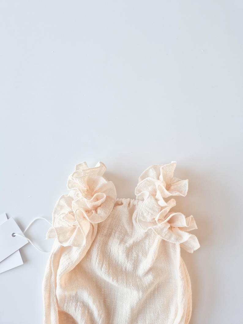 Detailed top-down view of an oversized frill collar on a baby girl romper. Creamy off-white textured cotton designed to drape beautifully over shoulders. Ideal baptism gift girl offering timeless elegance and a nostalgic glow.