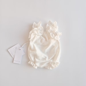 Classic presentation of an ivory baby romper laying flat with product tags. The non-reflective cotton material and elegant ruffle silhouette make it a sophisticated baptism gift girl option for milestone celebrations.