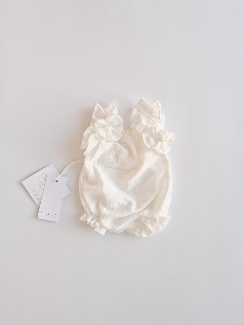 Classic presentation of an ivory baby romper laying flat with product tags. The non-reflective cotton material and elegant ruffle silhouette make it a sophisticated baptism gift girl option for milestone celebrations.