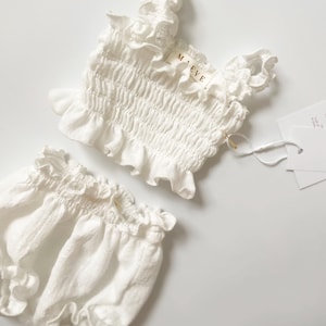 Flat lay studio photography of a white smocked two-piece set with a branded clothing tag. The macro shot reveals the deeply crinkled, tactile organic cotton weave and ruffled bodice, making it a luxurious, heirloom-quality newborn gift.