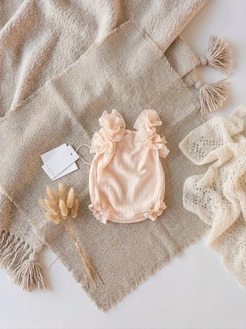 Flat lay styling of elegant baby girl clothes on a tactile linen cloth. This vanilla newborn outfit highlights breathable textured cotton and oversized frills. A camera-ready newborn gift or beautiful baptism gift girl selection.
