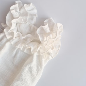 Macro shot of a baby girl clothes neckline focusing on the layered, off-white cotton ruffles. The high-volume frills create an organic, 3D structure designed to flatter delicate skin in newborn props photography.