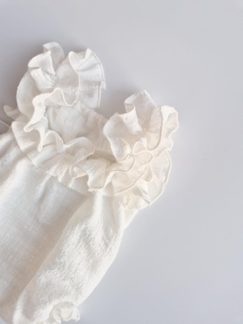 Macro shot of a baby girl clothes neckline focusing on the layered, off-white cotton ruffles. The high-volume frills create an organic, 3D structure designed to flatter delicate skin in newborn props photography.