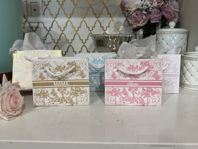 May include: Four small gift bags with white rope handles. The bags have a white background with a floral pattern and a gold or pink stripe. The bags are labelled "Mama" in gold and pink lettering.