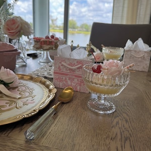 May include: A table setting featuring a pink and gold decorative plate, a gold spoon, a glass of pink drink with a cherry garnish, and a pink gift bag with the word "Alice" printed on it.