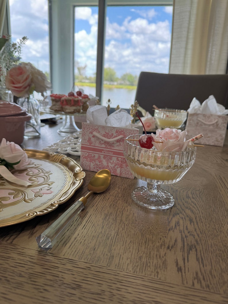 May include: A table setting featuring a pink and gold decorative plate, a gold spoon, a glass of pink drink with a cherry garnish, and a pink gift bag with the word "Alice" printed on it.