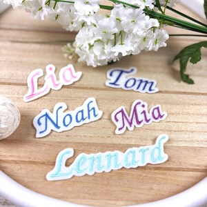 Könnte beinhalten: Fünf weiße Stoffaufnäher mit gestickten Namen in verschiedenen Farben. Die Namen Lia, Tom, Noah, Mia und Lennard sind in rosa, dunkelblauem, blauem, violettem und hellblauem Garn gestickt. Jeder Aufnäher hat einen weißen Rand.