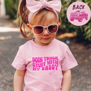 May include: A young child wearing a pink t-shirt with the text "DOIN TRUCK STUFF WITH MY DADDY" in a bubble font. The child is also wearing pink sunglasses and a pink bow headband. A pink circle with a truck graphic is visible.