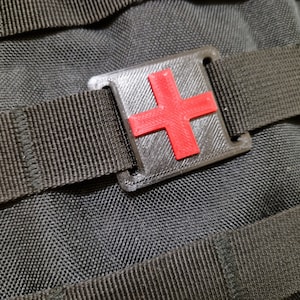 Könnte beinhalten: Eine schwarze Nylontasche mit einem roten medizinischen Kreuz, das auf eine schwarze Kunststoffschnalle gedruckt ist.